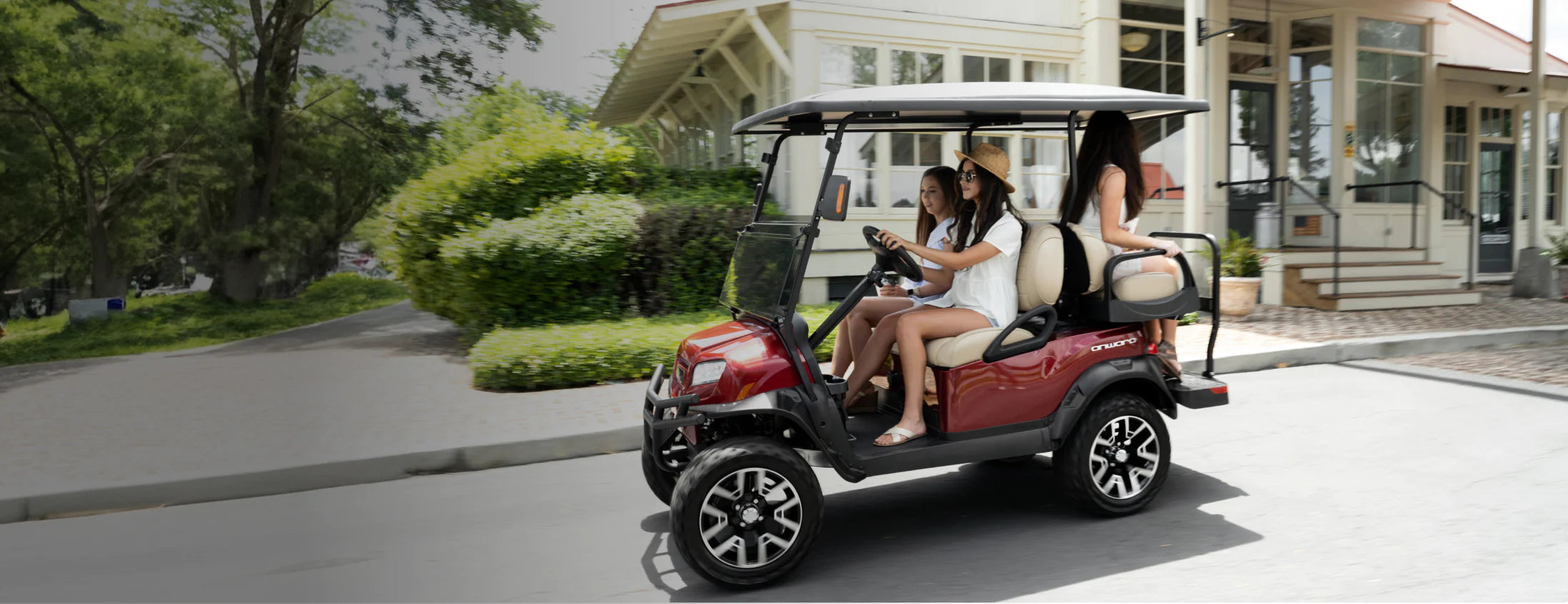 Three women ride on a dark red golf cart with oversized tires and a full-sized roof as they pass a store on a quiet street.